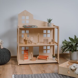 Puede incluir: Una casa de mu&ntilde;ecas de madera de tres pisos con techo a dos aguas. La casa de mu&ntilde;ecas est&aacute; amueblada con muebles en miniatura, incluyendo un sof&aacute;, sillas y una cama. Un cordero de peluche est&aacute; sentado en un puf gris. Una caja de madera contiene un animal de peluche.