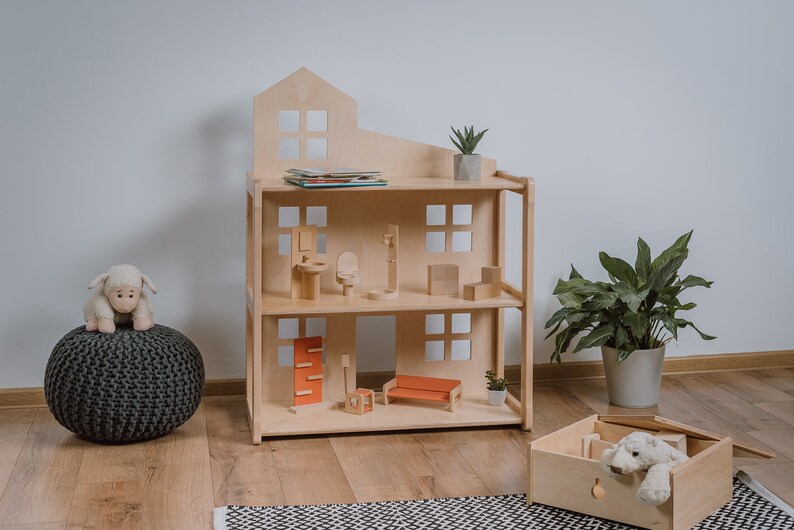 Puede incluir: Una casa de mu&ntilde;ecas de madera de tres pisos con techo a dos aguas. La casa de mu&ntilde;ecas est&aacute; amueblada con muebles en miniatura, incluyendo un sof&aacute;, sillas y una cama. Un cordero de peluche est&aacute; sentado en un puf gris. Una caja de madera contiene un animal de peluche.
