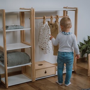 K&ouml;nnte beinhalten: Ein wei&szlig;er Kinderkleiderschrank aus Holz mit Regalen und einer Kleiderstange. Ein Kind steht vor dem Schrank und betrachtet die Kleidung. An der Stange h&auml;ngen zwei Hemden und eine Hose.