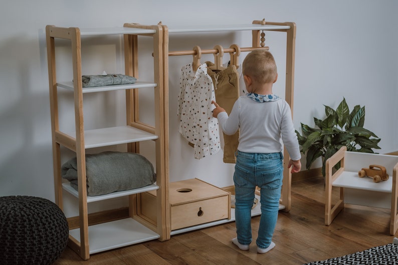 K&ouml;nnte beinhalten: Ein wei&szlig;er Kinderkleiderschrank aus Holz mit Regalen und einer Kleiderstange. Ein Kind steht vor dem Schrank und betrachtet die Kleidung. An der Stange h&auml;ngen zwei Hemden und eine Hose.