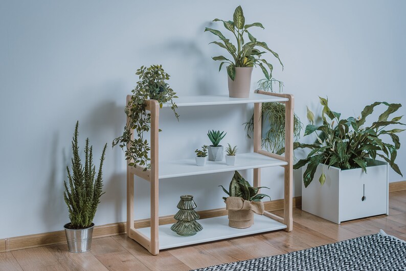 Puede incluir: Una estanter&iacute;a de madera de tres niveles con estantes blancos y soportes de madera natural. La estanter&iacute;a est&aacute; llena de varias plantas en macetas, incluyendo suculentas, hojas verdes y enredaderas colgantes. Tambi&eacute;n se exhiben un &aacute;rbol decorativo de cristal y una maceta envuelta en arpillera.