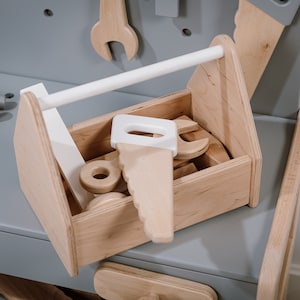 May include: A wooden toy toolbox with a white handle, filled with various wooden tools, including a saw, wrench, and other tools. The toolbox is sitting on a gray surface.