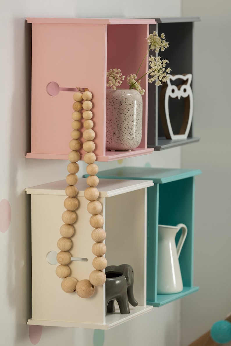May include: Four square wooden shelves in pink, grey, white, and teal with decorative items on each shelf. The top shelf has a small vase with white flowers and a white owl figurine. The second shelf has a wooden bead necklace. The third shelf has a small grey elephant figurine. The bottom shelf has a white jug.