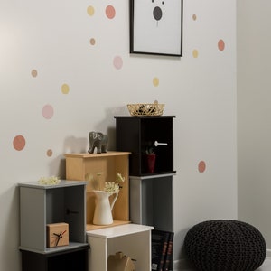May include: A set of black, white, grey, and wood cube shelves with various decorative items, including a small elephant figurine, a white jug with flowers, a clock, a vase, and a small house figurine. The shelves are against a white wall with pink, yellow, and orange polka dots.