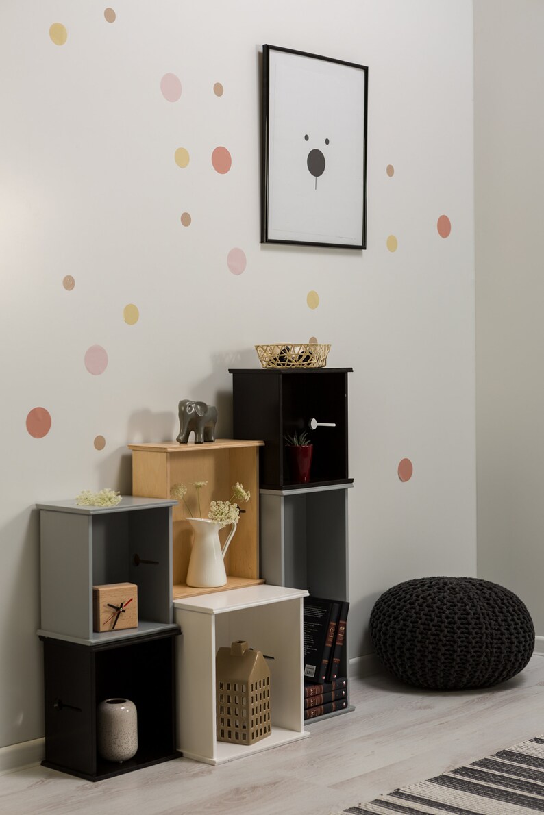 May include: A set of black, white, grey, and wood cube shelves with various decorative items, including a small elephant figurine, a white jug with flowers, a clock, a vase, and a small house figurine. The shelves are against a white wall with pink, yellow, and orange polka dots.