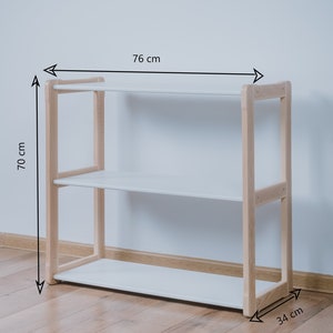 May include: White shelf unit with three shelves and a natural wood frame. The shelf unit is 76 cm wide, 70 cm tall, and 34 cm deep.