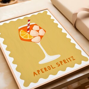 Peut inclure: Une impression encadr&eacute;e avec un fond jaune repr&eacute;sentant un verre d'Aperol Spritz avec une tranche d'orange et des gla&ccedil;ons. Le texte "APEROL SPRITZ" est &eacute;crit en lettres orange en bas de l'impression.