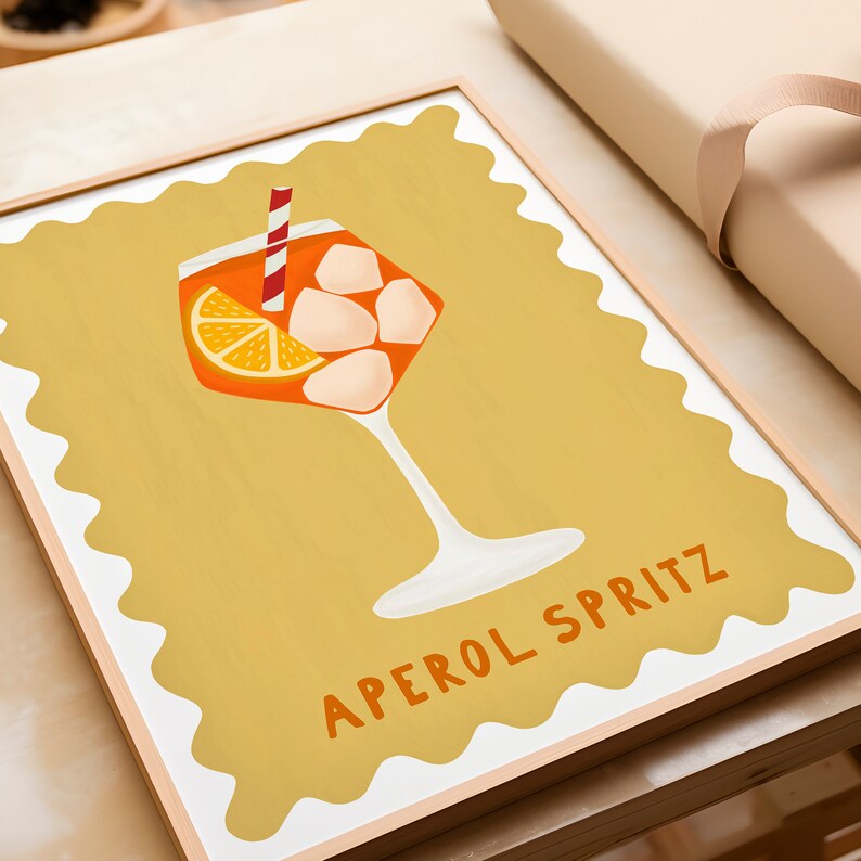 Peut inclure: Une impression encadr&eacute;e avec un fond jaune repr&eacute;sentant un verre d'Aperol Spritz avec une tranche d'orange et des gla&ccedil;ons. Le texte "APEROL SPRITZ" est &eacute;crit en lettres orange en bas de l'impression.