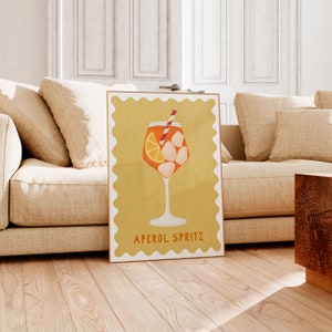Peut inclure: Une affiche jaune avec une bordure blanche et un bord festonn&eacute;. L'affiche repr&eacute;sente un verre d'Aperol Spritz avec une tranche d'orange, des gla&ccedil;ons et une paille rouge et blanche. Le texte "APEROL SPRITZ" est imprim&eacute; en noir en bas de l'affiche.