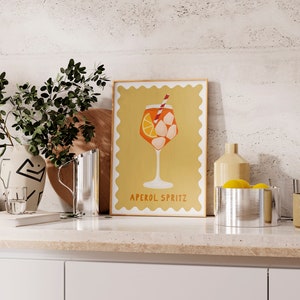 Peut inclure: Une illustration num&eacute;rique d'un cocktail Aperol Spritz dans un verre avec une paille et des gla&ccedil;ons. L'illustration est sur un fond jaune avec une bordure blanche et le texte "Aperol Spritz" en bas.