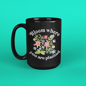 May include: Black ceramic mug with a curved handle. The mug features a floral design in pink, blue, and yellow, with the text "Bloom where you are planted" in a decorative font. The mug is set against a teal background.