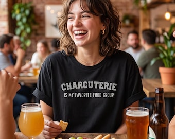 May include: A black t-shirt with the white text "CHARCUTERIE IS MY FAVORITE FOOD GROUP" printed on the front. The shirt features a crew neckline and short sleeves, worn by a person sitting at a wooden table with a charcuterie board and beer.