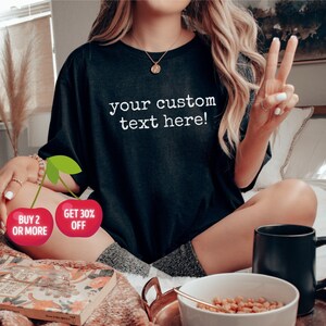 Puede incluir: Una camiseta negra con el texto "your custom text here!" impreso en blanco. La camiseta la lleva puesta una persona sentada en una cama. Hay dos cerezas rojas con texto blanco que dicen "Buy 2 or More" y "Get 30% Off".