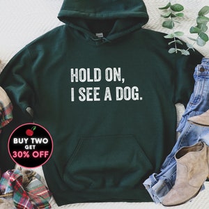 Pode incluir: Uma camisa de moletom com capuz verde escura com o texto "HOLD ON, I SEE A DOG." impresso em branco na frente.