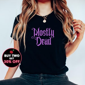 May include: A black t-shirt with the text "Mostly Dead" in purple script. The shirt is being worn by a person with long blonde hair and is sitting on a red chair. The text "BUY TWO GET 30% OFF" is in a red circle with a cherry graphic.