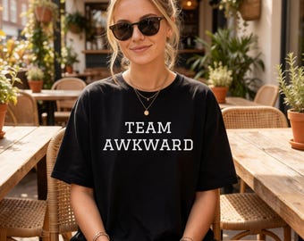 Puede incluir: Una camiseta negra de manga corta con el texto blanco TEAM AWKWARD impreso en una fuente sans-serif en negrita en el pecho. La prenda tiene un cuello redondo clásico y un corte relajado, ofreciendo un diseño sencillo y directo.