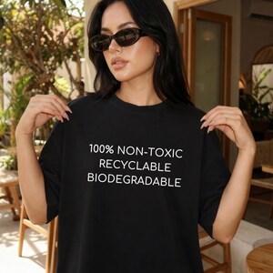 May include: Black t-shirt with white text: "100% NON-TOXIC, RECYCLABLE, BIODEGRADABLE." The shirt is a solid black color. The model is wearing sunglasses. The shirt is a casual style.