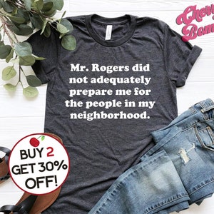 Puede incluir: Una camiseta gris oscuro con texto blanco que dice "Mr. Rogers did not adequately prepare me for the people in my neighborhood." La camiseta est&aacute; doblada y se muestra sobre una superficie blanca con un par de jeans azules y un gr&aacute;fico de cereza roja con el texto "BUY 2 GET 30% OFF!"
