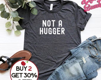 May include: A dark grey t-shirt with the text "NOT A HUGGER" in white. The shirt is laid flat on a white surface with a pair of brown sandals and blue jeans. A red circle with a white cherry and the text "BUY 2 GET 30% OFF!" is in the bottom left corner.