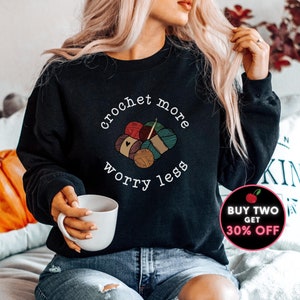 May include: A black sweatshirt with the text "crochet more worry less" in white lettering. The text is surrounded by a graphic of colorful yarn balls and knitting needles.