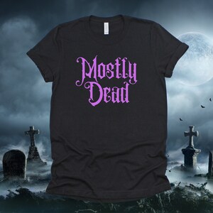 May include: Black t-shirt with a purple graphic that says "Mostly Dead" in a gothic font. The shirt is photographed against a dark background with a full moon and two crosses.