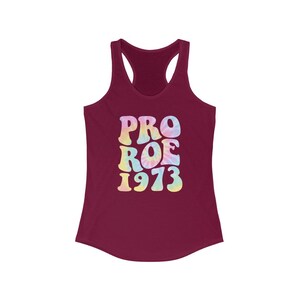May include: A burgundy racerback tank top with the words "PRO ROE 1973" in a tie-dye design. The text is in a retro font with pastel colors, including pink, yellow, and blue. The tank top has a scoop neck and a sleeveless design.