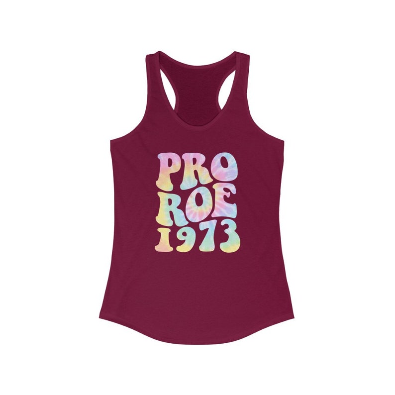 May include: A burgundy racerback tank top with the words "PRO ROE 1973" in a tie-dye design. The text is in a retro font with pastel colors, including pink, yellow, and blue. The tank top has a scoop neck and a sleeveless design.