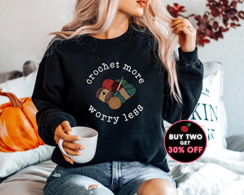 May include: A black sweatshirt with the text "crochet more worry less" in white lettering. The text is surrounded by a graphic of colorful yarn balls and knitting needles.