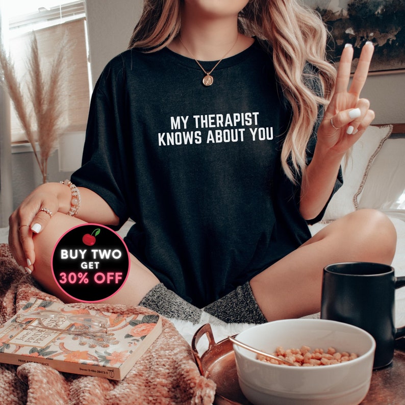 May include: A black t-shirt with the text "My therapist knows about you" in white. The t-shirt is being worn by a person sitting on a bed. A red and white circle with the text "Buy two get 30% off" is in the lower left corner of the image.
