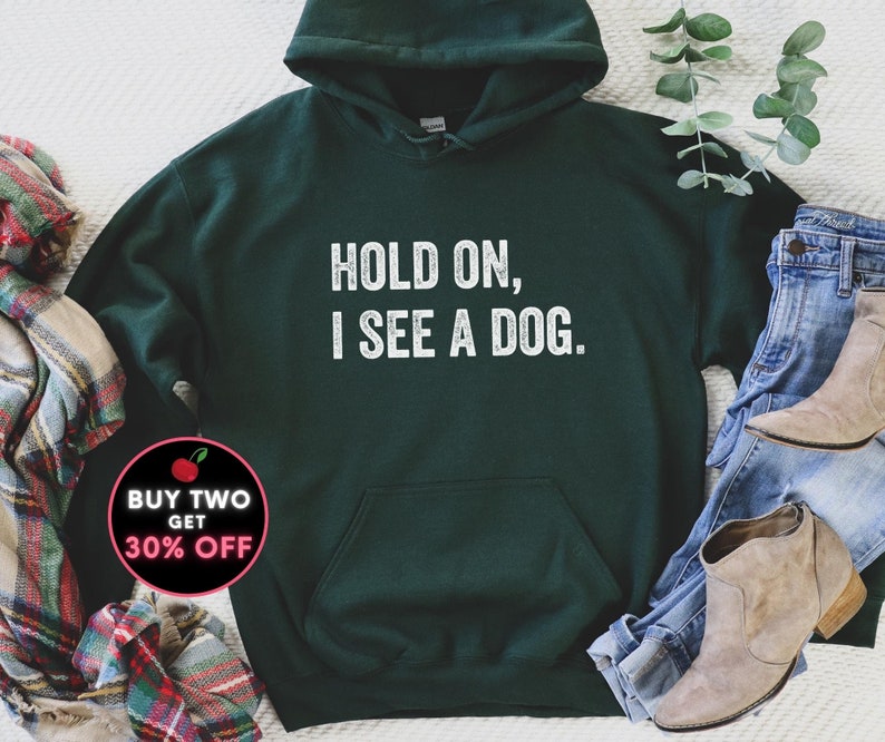 Pode incluir: Uma camisa de moletom com capuz verde escura com o texto "HOLD ON, I SEE A DOG." impresso em branco na frente.
