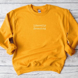 May include: Mustard yellow sweatshirt with the phrase "literally freezing" in white, centered on the chest. The long-sleeved crew neck top is made of a soft material. The Gildan brand tag is visible on the back of the neck.