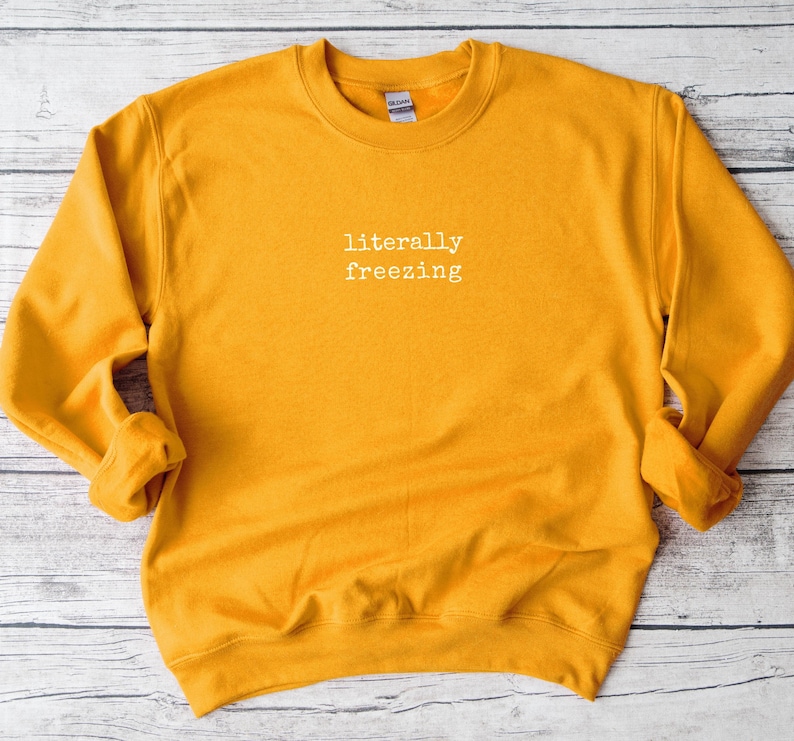 May include: Mustard yellow sweatshirt with the phrase "literally freezing" in white, centered on the chest. The long-sleeved crew neck top is made of a soft material. The Gildan brand tag is visible on the back of the neck.
