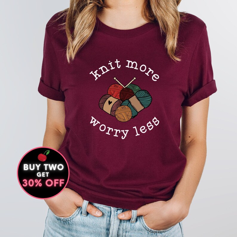 May include: A maroon t-shirt with rolled-up sleeves featuring the text "knit more worry less" in white. The graphic includes yarn balls and knitting needles. A circular promotion graphic is visible in the lower left corner.