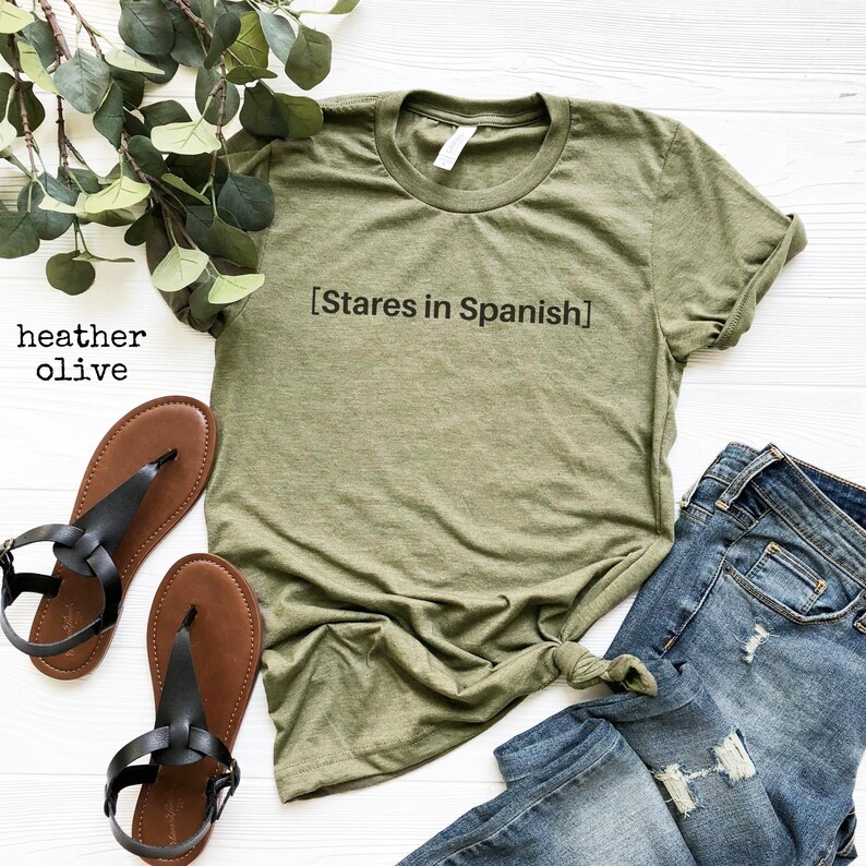 以下が含まれることがあります： 前面に「[Stares in Spanish]」という文字が印刷された緑色のTシャツ。白い背景に、茶色のサンダルと青いジーンズと一緒に置かれています。