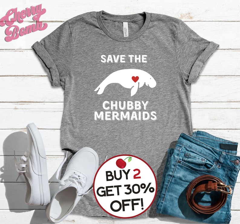 May include: Gray t-shirt with white text that says "Save the Chubby Mermaids" and a white silhouette of a manatee with a red heart. The t-shirt is on a white wood background with a pair of white sneakers and blue jeans. A red circle with a white cherry and white text that says "Buy 2 Get 30% Off!" is in the lower right corner.