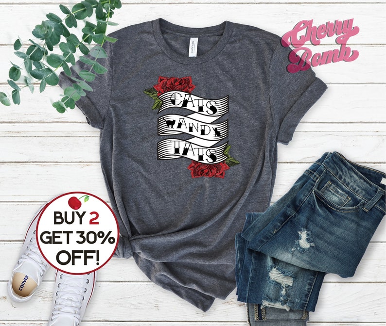 May include: A heather grey t-shirt with a white graphic that says "Cats and Tats" with a red rose design. The shirt is displayed on a white wooden surface with a pair of white trainers and blue jeans.