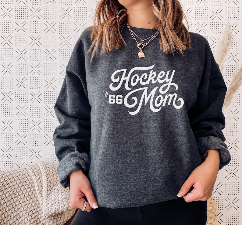 Może przedstawiać: Szara bluza z kapturem z napisem "Hockey #66 Mom" w kolorze białym. Bluza ma luźny kr&oacute;j i okrągły dekolt.