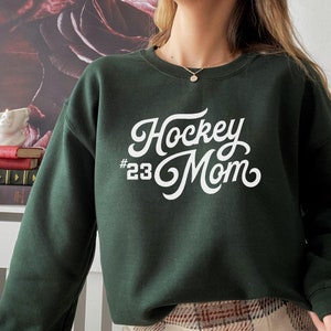 K&ouml;nnte beinhalten: Ein dunkelgr&uuml;ner Pullover mit wei&szlig;em Text, der "Hockey #23 Mom" in einer Retro-Schriftart lautet.