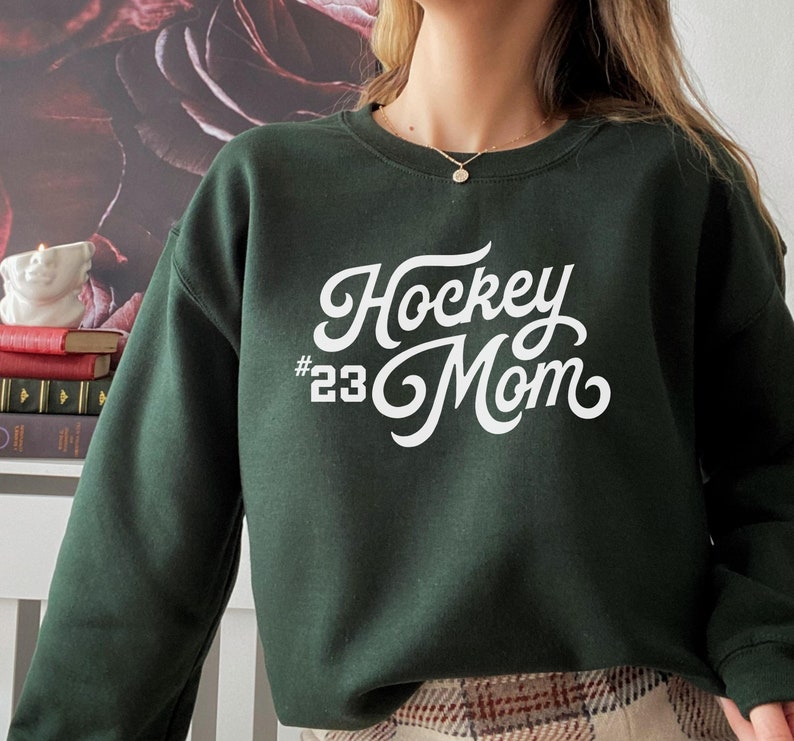 K&ouml;nnte beinhalten: Ein dunkelgr&uuml;ner Pullover mit wei&szlig;em Text, der "Hockey #23 Mom" in einer Retro-Schriftart lautet.