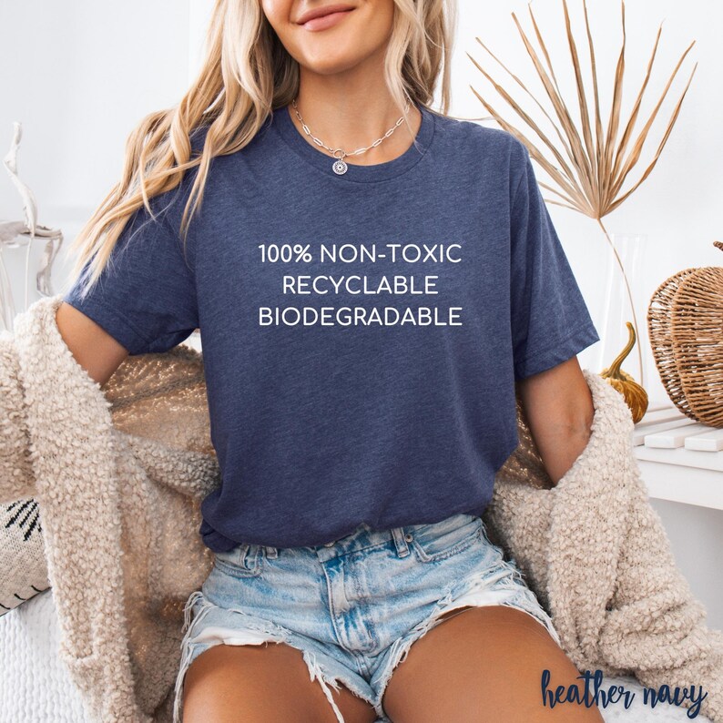 May include: Heather navy blue t-shirt with white text that reads "100% NON-TOXIC RECYCLABLE BIODEGRADABLE." The shirt is short-sleeved and has a crew neck. The model is wearing denim shorts and a cream-colored cardigan.