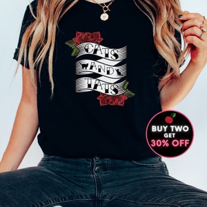May include: Black t-shirt featuring a white graphic that reads "Cats and Tats" with a red rose design. The graphic is on a banner with a black and white striped pattern. The t-shirt is being worn by a person with long blonde hair. There is a red cherry with the text "Buy Two Get 30% Off" in a white circle.