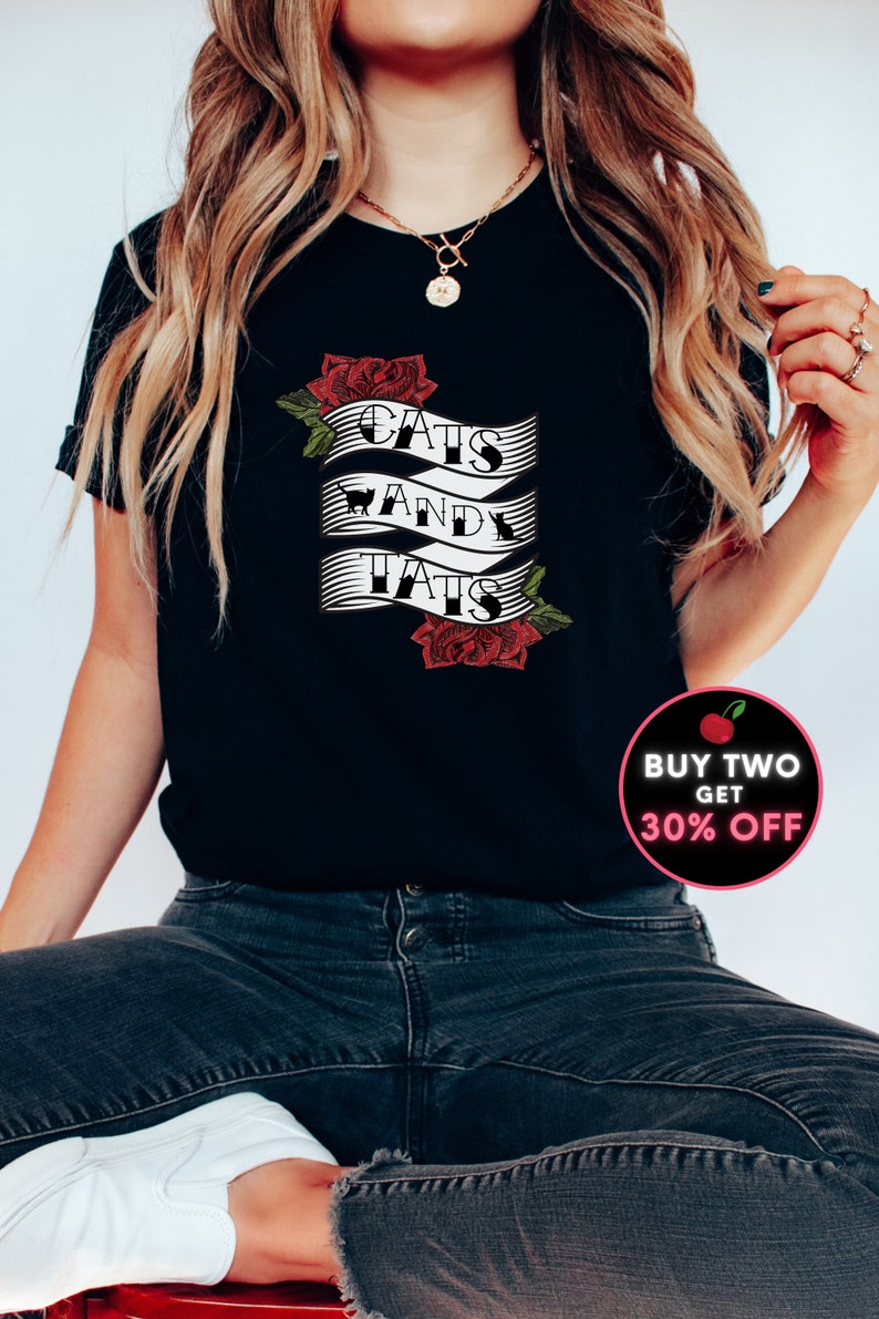 May include: Black t-shirt featuring a white graphic that reads "Cats and Tats" with a red rose design. The graphic is on a banner with a black and white striped pattern. The t-shirt is being worn by a person with long blonde hair. There is a red cherry with the text "Buy Two Get 30% Off" in a white circle.