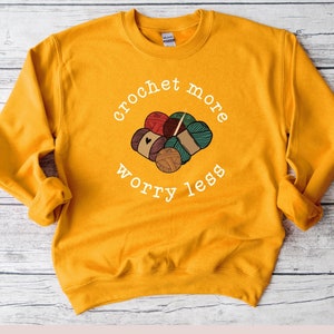 May include: A mustard yellow sweatshirt with the text "crochet more worry less" in white. The text is surrounded by a circle and a graphic of yarn balls and knitting needles.