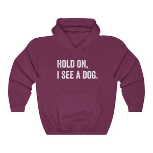 Pode incluir: Uma camisola de capuz cor vinho com o texto "HOLD ON, I SEE A DOG." impresso a branco na frente.