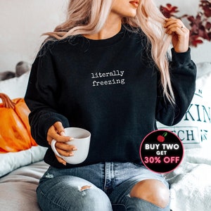May include: Black sweatshirt with the text "literally freezing" in white. The sweatshirt is a crew neck style. The image also includes a white mug and a promotional graphic.