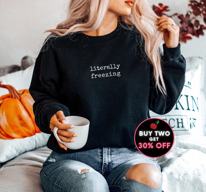 May include: Black sweatshirt with the text "literally freezing" in white. The sweatshirt is a crew neck style. The image also includes a white mug and a promotional graphic.