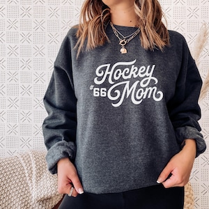 K&ouml;nnte beinhalten: Ein dunkelgrauer Pullover mit dem Text "Hockey #66 Mom" in wei&szlig;er Kursivschrift.