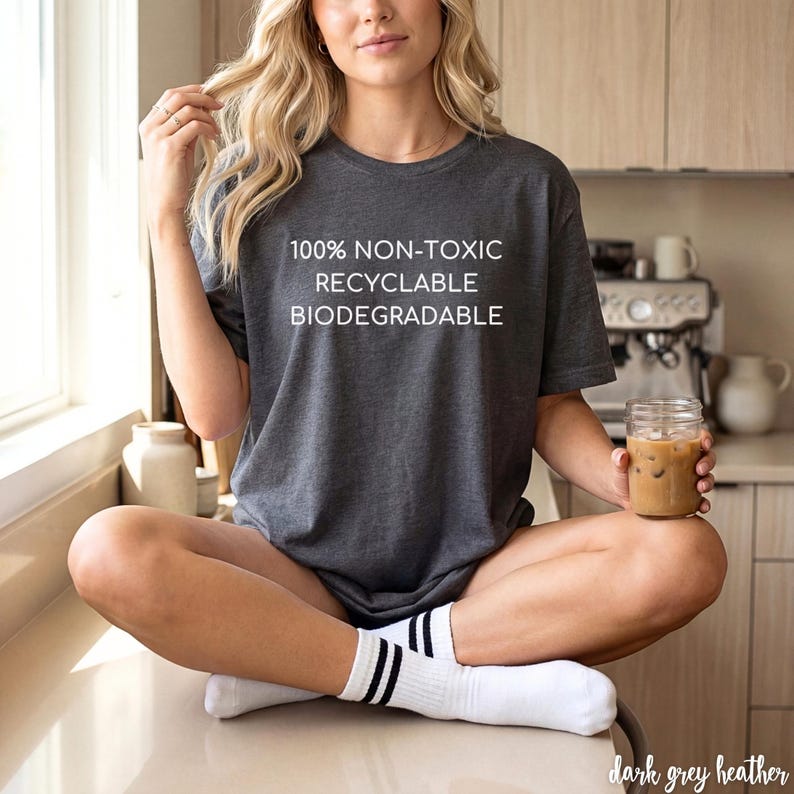 May include: Dark grey heather t-shirt with the text "100% NON-TOXIC RECYCLABLE BIODEGRADABLE". The shirt is paired with white socks with black stripes. A glass jar with a drink is held in the hand.