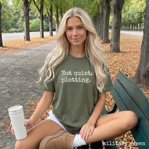 May include: Military green t-shirt with the text "Not quiet; plotting." in white. The shirt is a crew neck style. A white travel mug is held in the left hand. The model is sitting on a park bench.