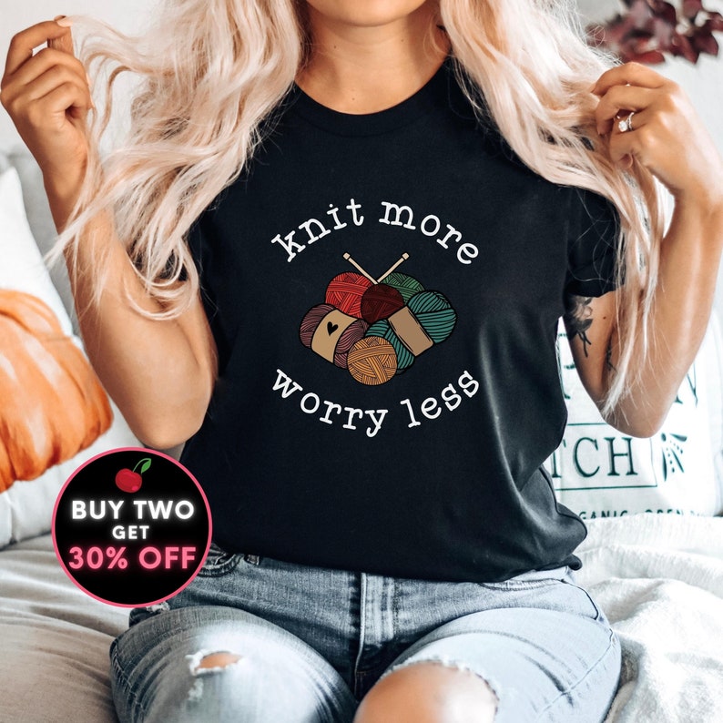 May include: Black t-shirt with the text "knit more worry less" above a graphic of yarn balls and knitting needles. The yarn balls are in shades of red, green, brown, and tan. The shirt is worn with blue jeans.